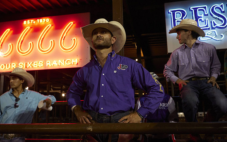 Man wearing a purple shirt and a cowboy hat standing in front of a big screen at a rodeo.